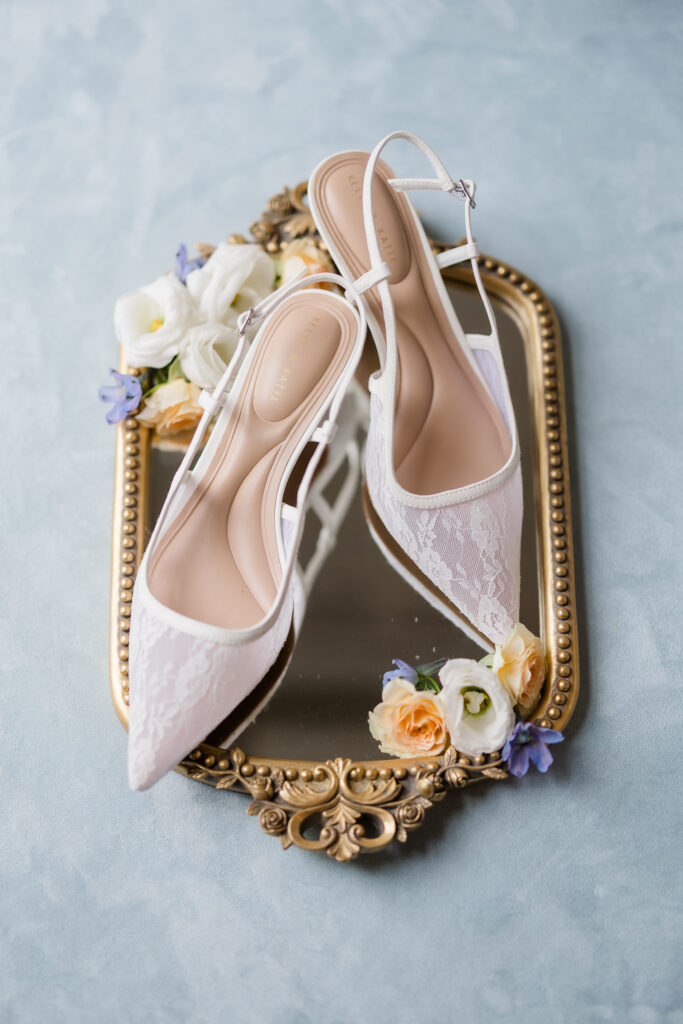 Elegant white lace wedding heels on gold vintage mirror surrounded by pastel flowers.