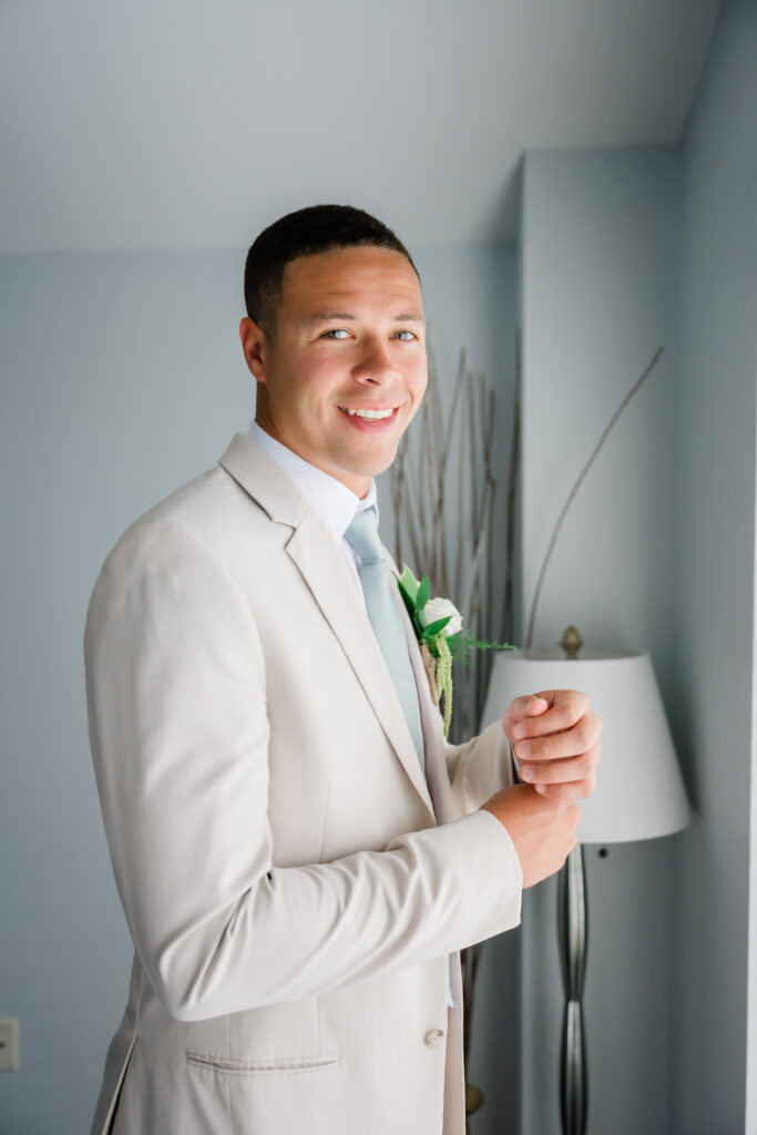 Groom in light beige suit and blue tie sitting in a modern chair, smiling before wedding ceremony at The Cathedral Basilica in Covington KY.