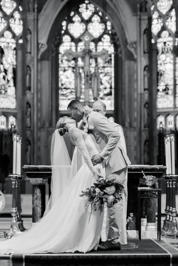 Bride and groom sharing first kiss in front of stained-glass altar at the Cathedral Basilica of the Assumption in Covington KY.